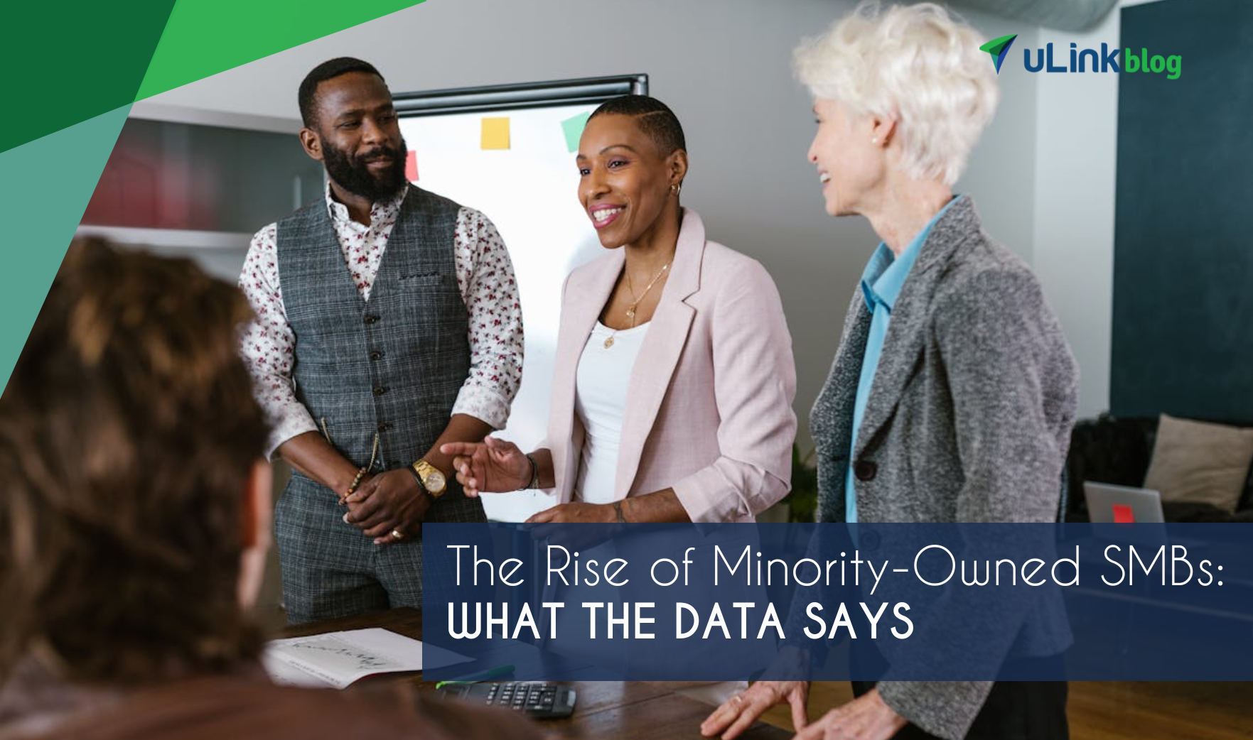 A Black man, Black woman, and older white woman stand in front of a desk giving a business presentation.